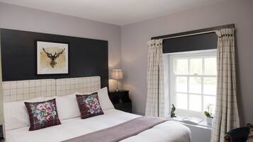 A bedroom with a large bed, a headboard with a grid pattern, a framed deer head artwork, a bedside lamp, a window with curtains, and a small table with a plant.