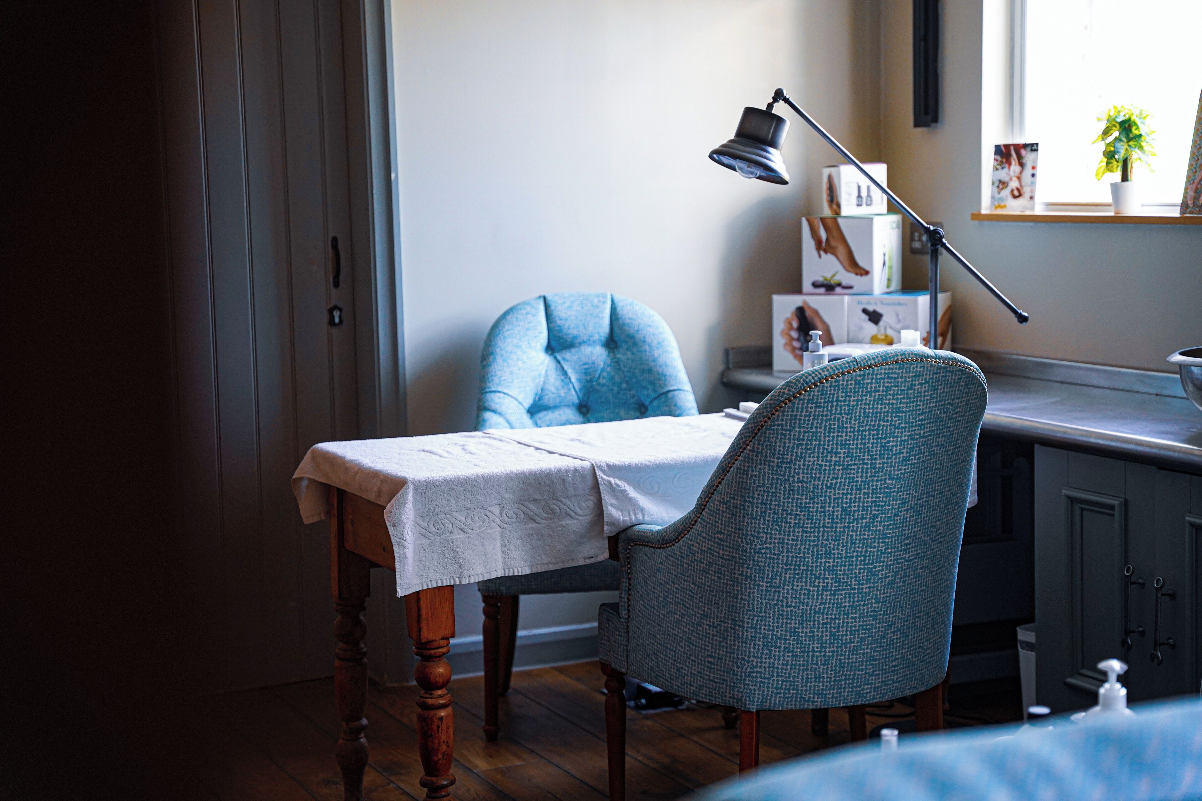 A room with a wooden table, two chairs, a desk, a lamp, and a window with a plant.