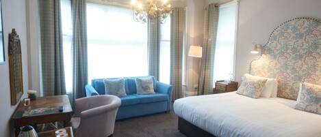 A hotel room with a large bed, a blue sofa, a chair, a desk with a laptop, and a chandelier.