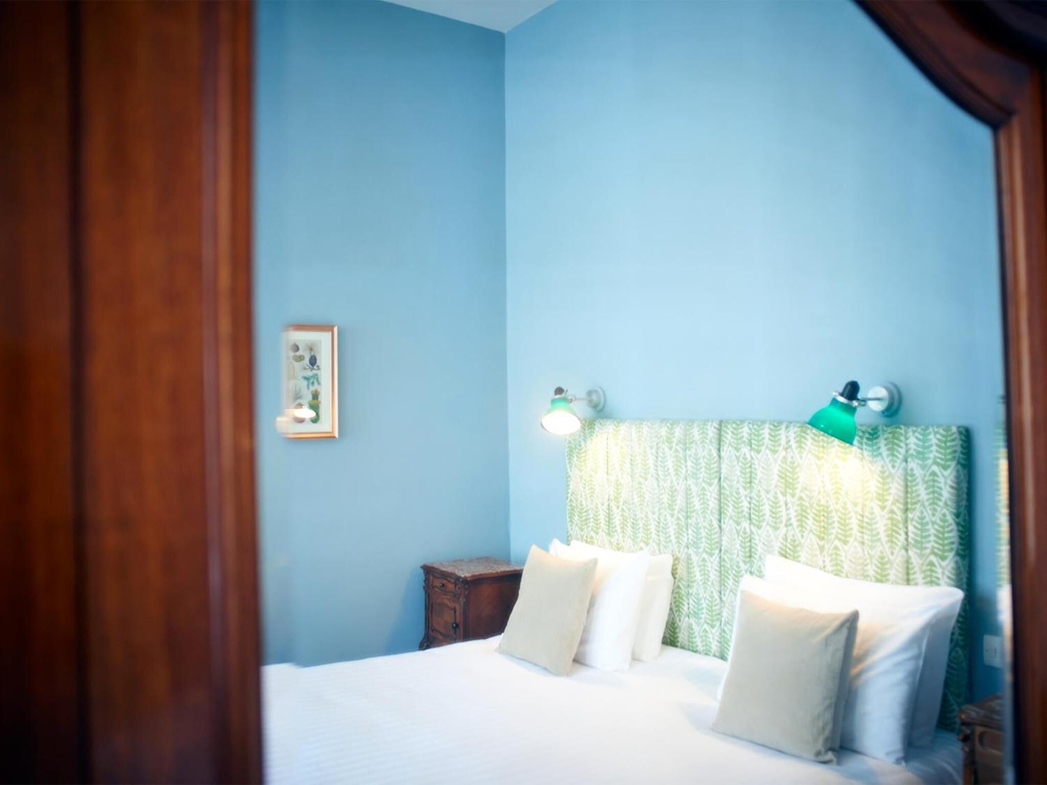 Un dormitorio con cama, almohadas, una mesita de noche, un cuadro enmarcado y lámparas fijadas a la pared.