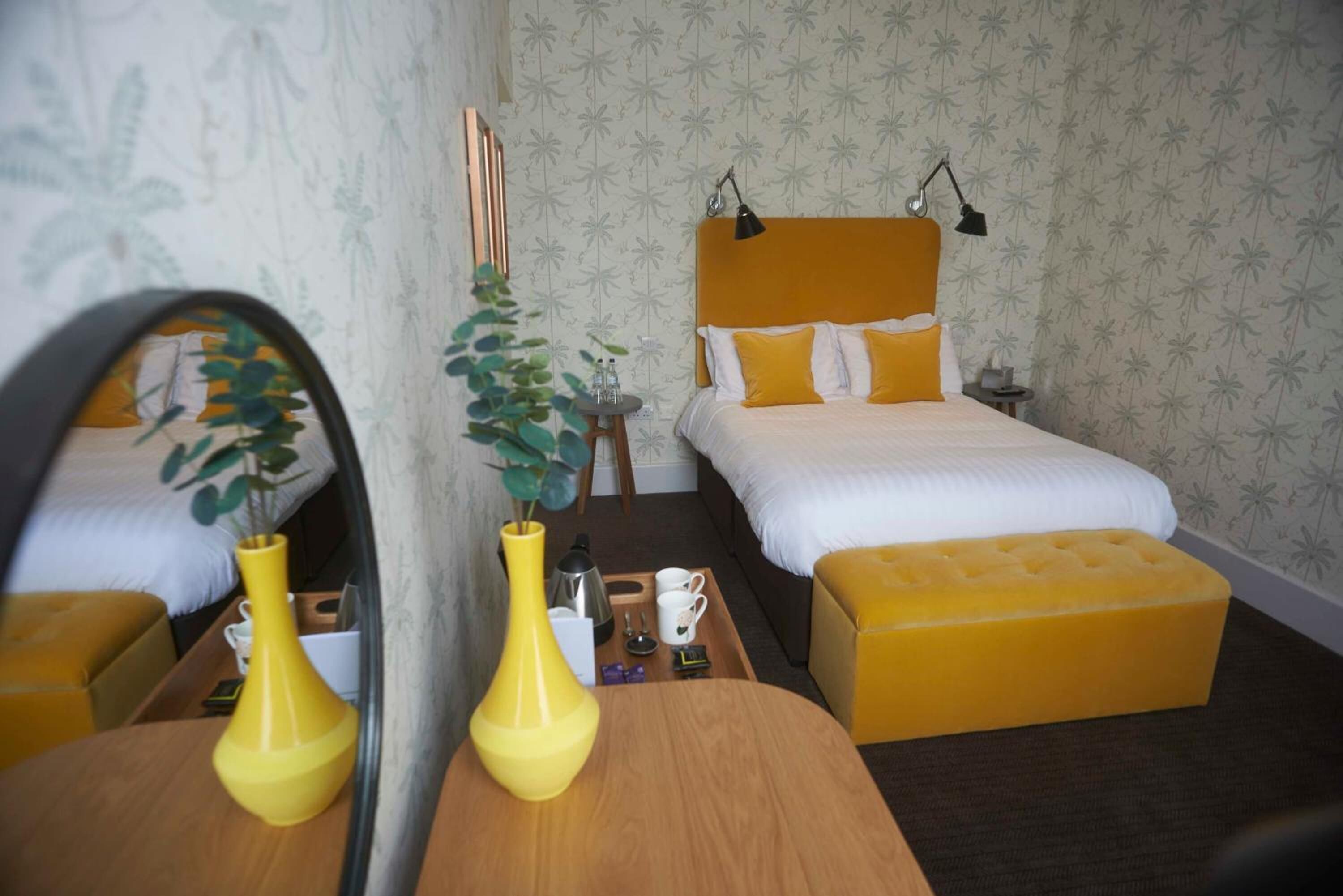 Habitación de hotel con dos camas, una mesita de noche de madera, un espejo y un jarrón amarillo con una planta.