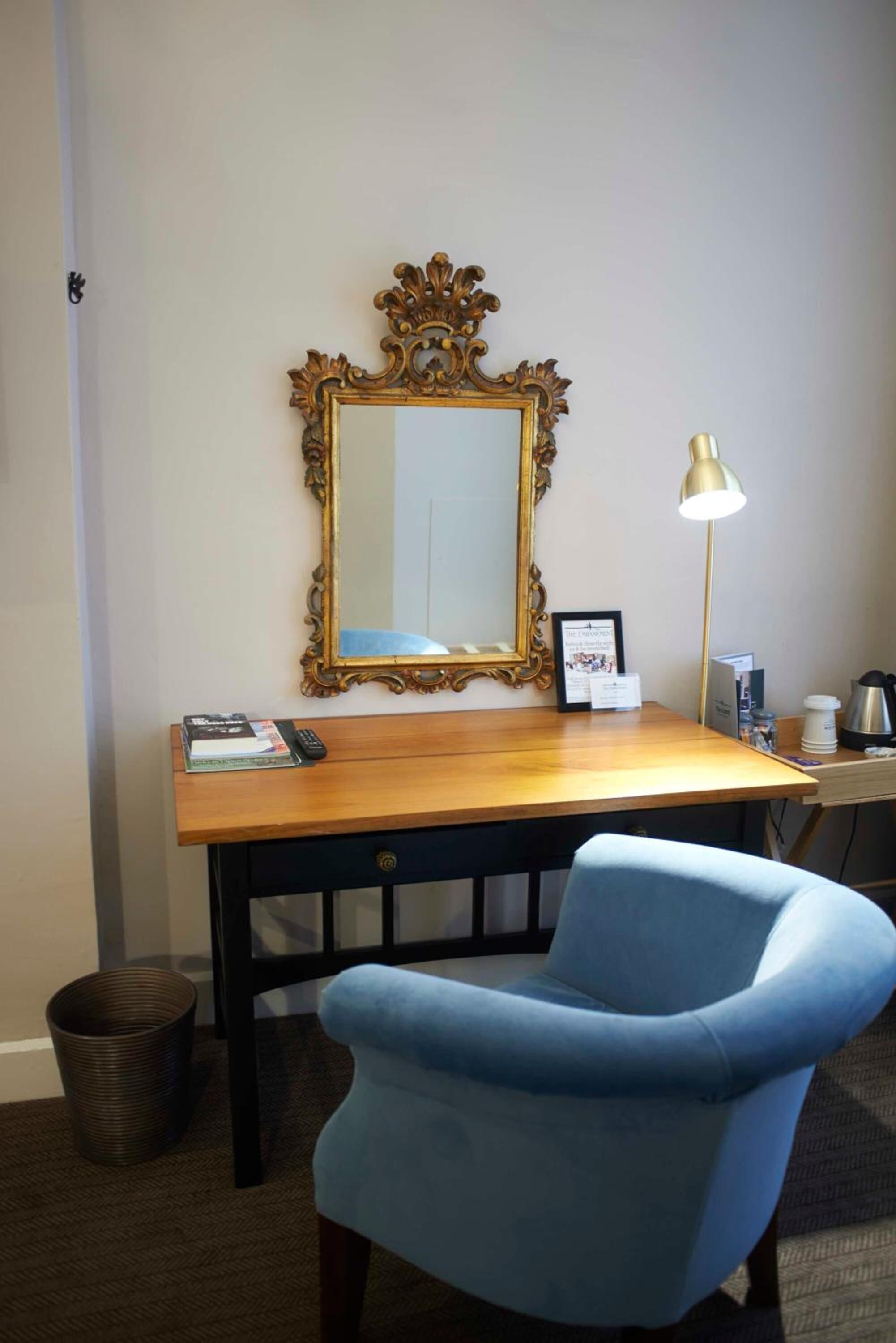A room with a wooden desk, a blue chair, a large ornate mirror, a lamp, and a trash bin.