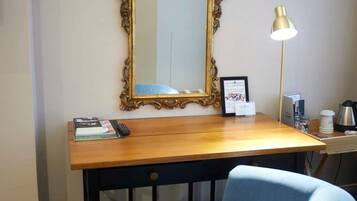 A room with a wooden desk, a blue chair, a large ornate mirror, a lamp, and a trash bin.