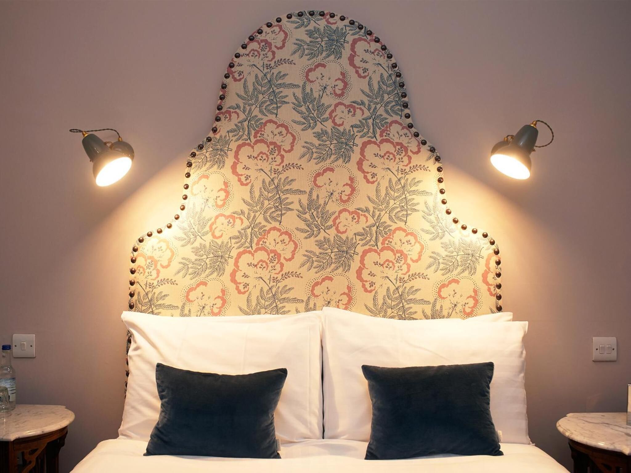 Una cama con sábanas blancas y dos almohadas oscuras, un cabecero estampado con motivos florales y dos lámparas fijadas a la pared.