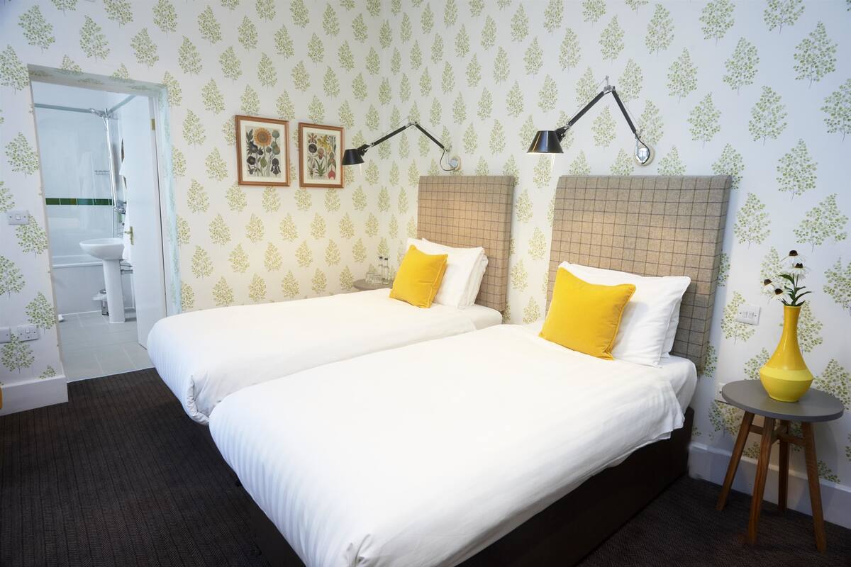A hotel room with two beds, a bathroom visible through an open door, and a small table with a vase.