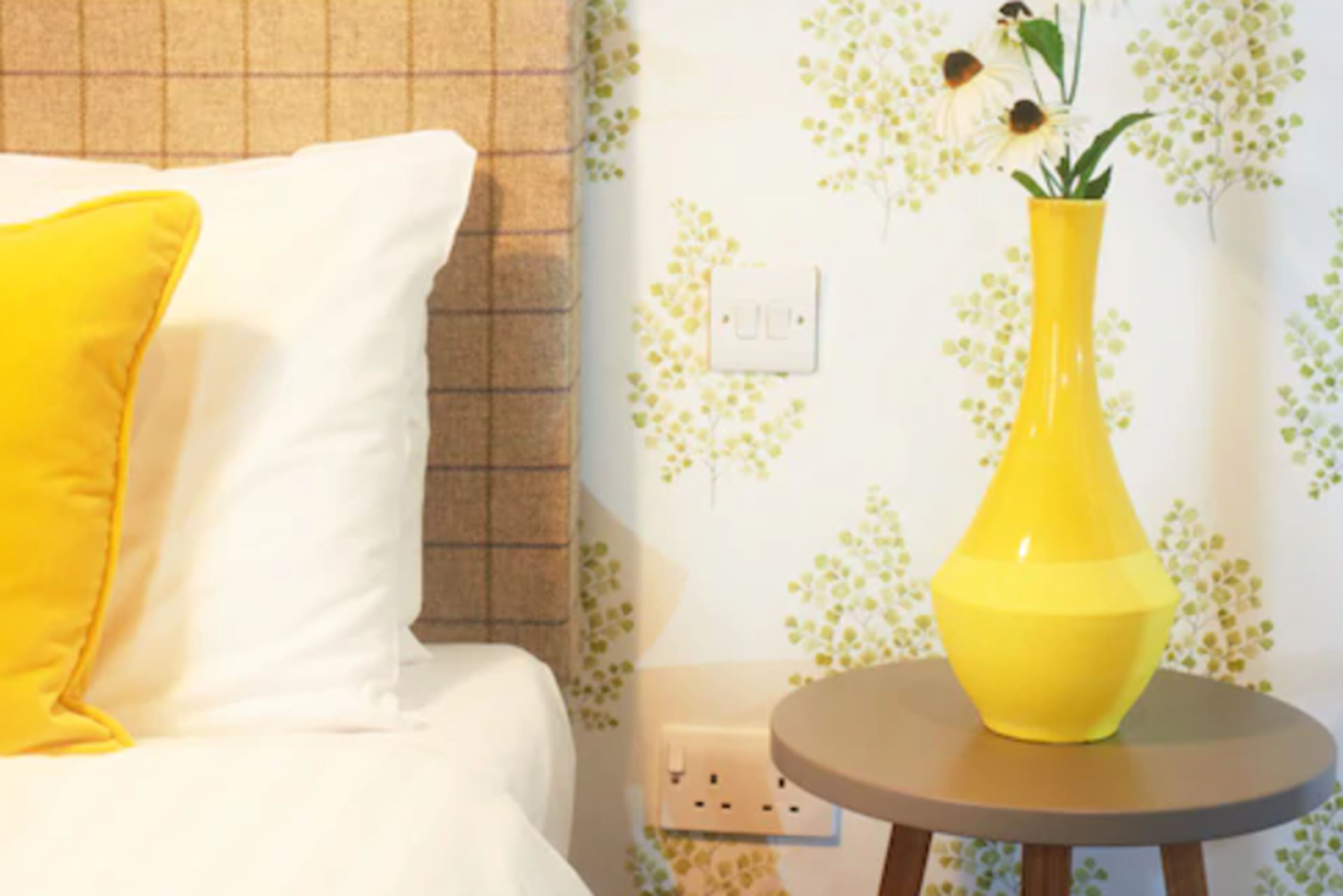 Una cama con un almohadón blanco y uno amarillo, un jarrón amarillo con flores sobre una mesita redonda, y una pared con estampado floral.