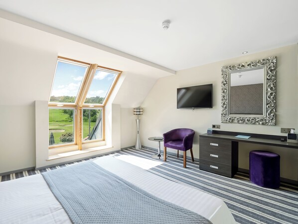 A bedroom with a large bed, a desk, a chair, a TV, and a window with a view of a green landscape. - Wharton Park Golf & Country Club (Bewdley)