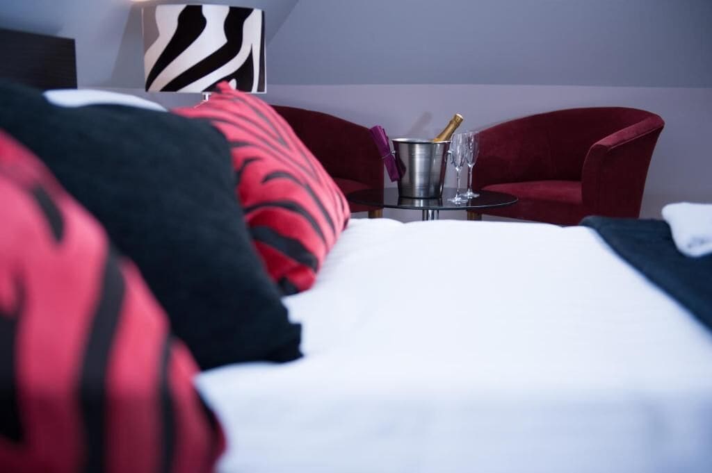 Quarto de hotel com uma cama, almofadas vermelhas e pretas, uma mesinha com uma garrafa e copos, e uma cadeira vermelha.