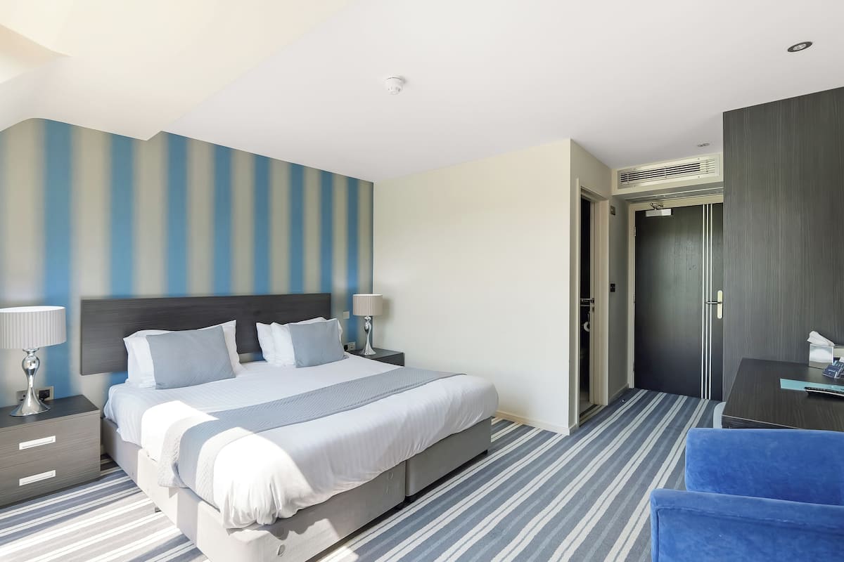 A modern hotel room with a large bed, a bedside table with a lamp, a blue chair, and a wardrobe.