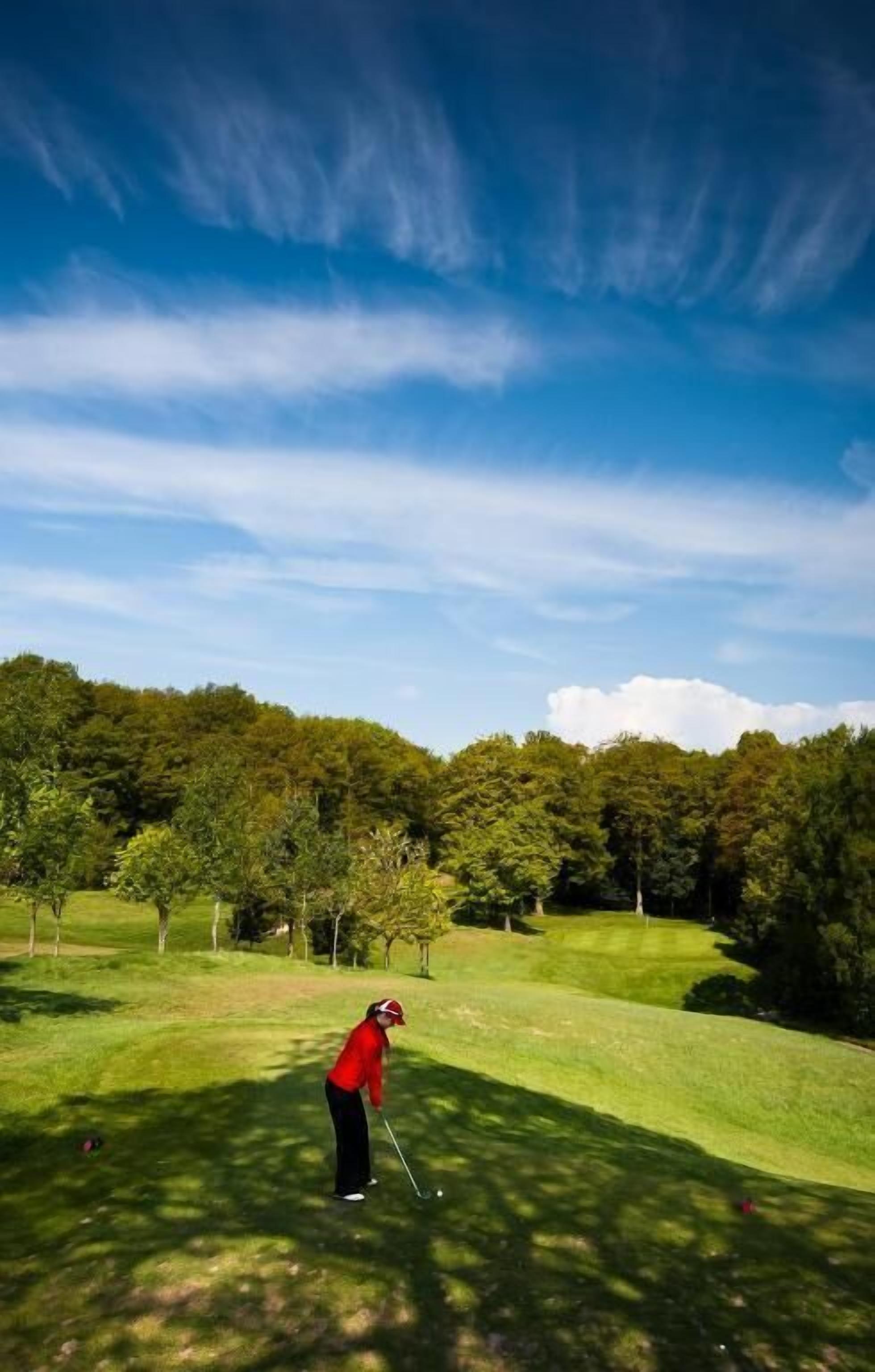 A person in a red jacket playing golf on a well-maintained course with lush greenery and a clear sky.