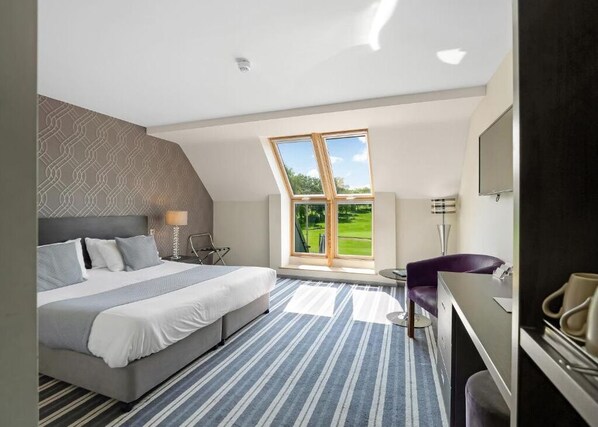 A modern hotel room with a large bed, a desk, and a view of a green landscape through a skylight. - Wharton Park Golf & Country Club (Bewdley)