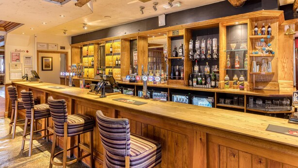 A bar area with wooden bar stools, a counter with a cash register, and shelves stocked with bottles. - Premier Inn Birmingham South - Rubery (Birmingham)