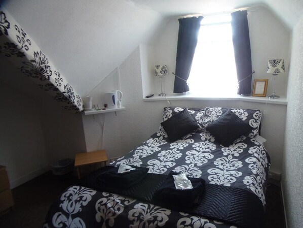 A small, neatly arranged bedroom with a bed, a nightstand, a shelf with a kettle, and a window with curtains. - Oban House Hotel (Blackpool)