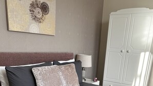 En pænt redt seng med mørkt sengetæppe og puder, en sengebordlampe og et klædeskab i baggrunden.
