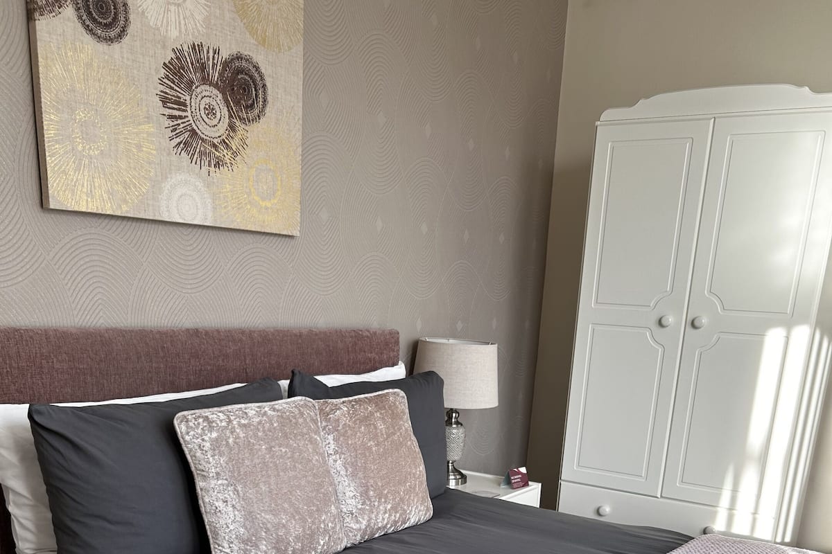 Un lit bien fait, avec une couvre-lit et des oreillers aux couleurs sombres, une lampe de chevet et une armoire en arrière-plan.