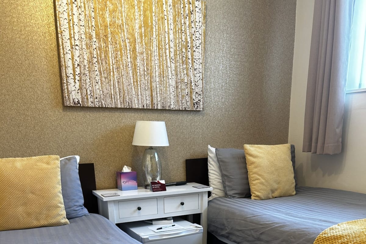 Une chambre d’hôtel avec deux lits, chacun recouvert d’une couverture jaune, une table de chevet avec une lampe, une boîte de mouchoirs et un panneau.