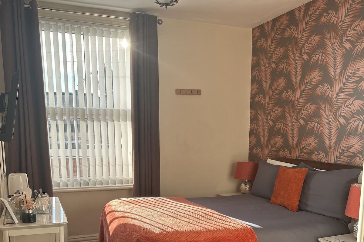 Une chambre à coucher comprenant un lit, une chaise, une table de chevet, une lampe, une fenêtre avec des rideaux et un mur orné d’un motif de feuilles.