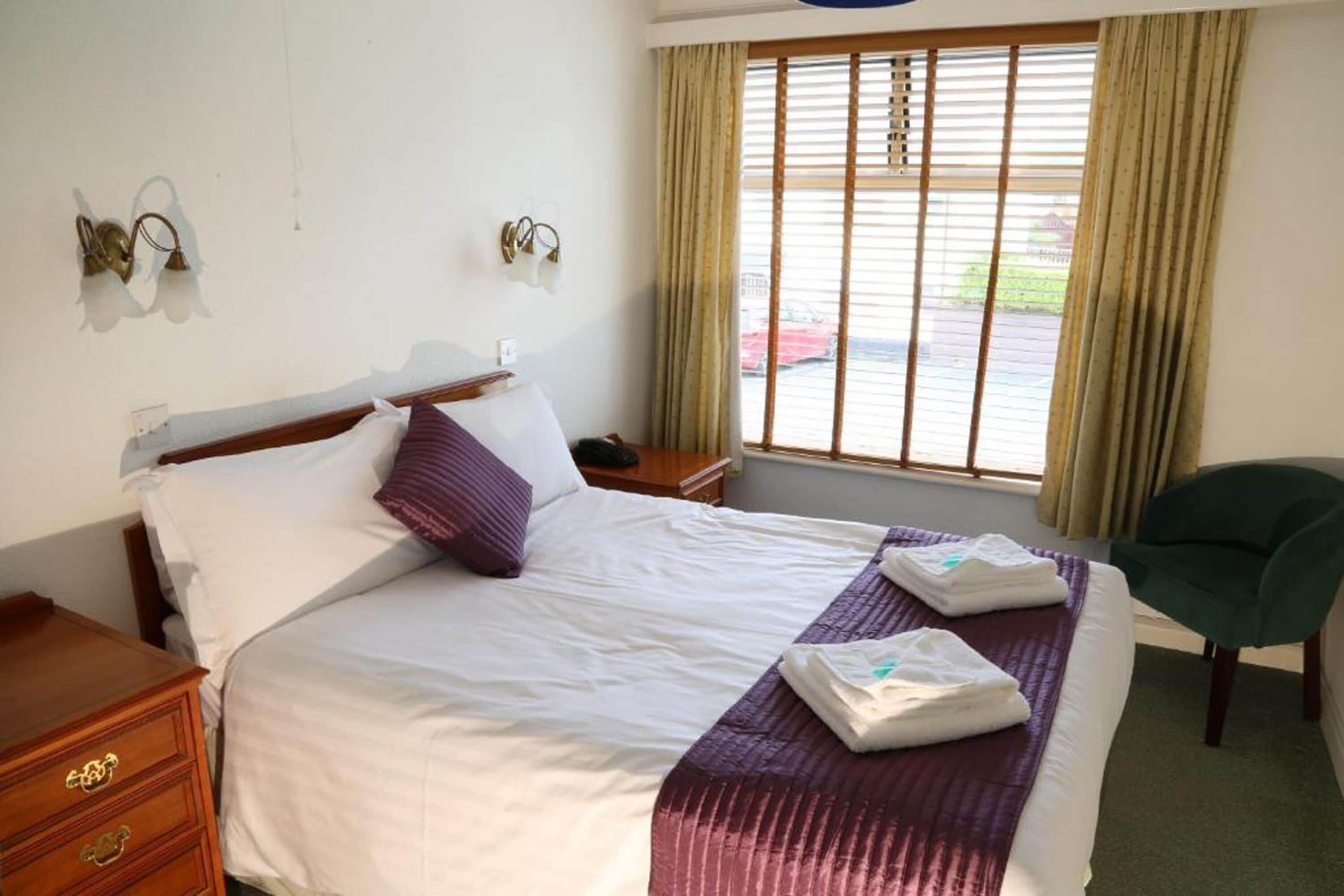 A neatly made bed with white and purple bedding, a wooden nightstand with drawers, a green chair, and a window with blinds.