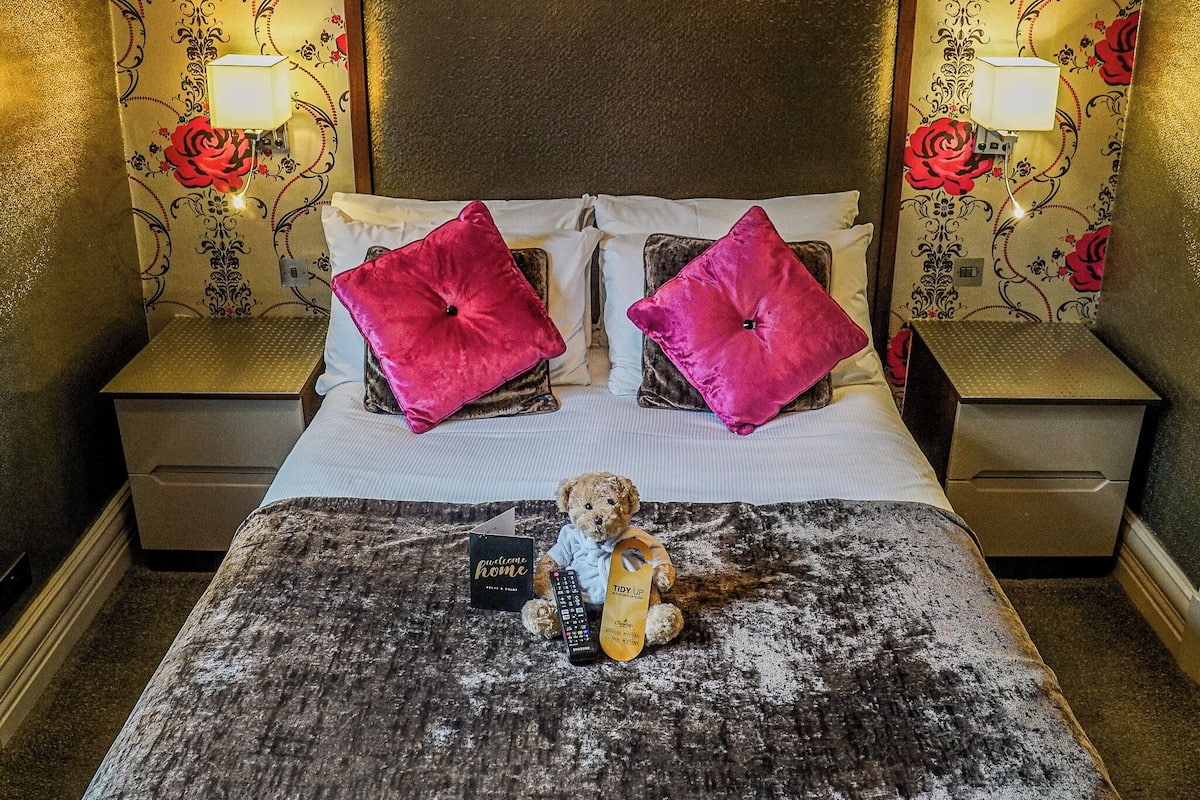 A neatly made bed with a teddy bear, two pillows, and a remote control on a textured blanket.