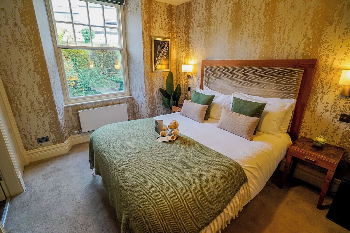 A bedroom with a large bed, a nightstand, a window with a view of greenery, and a wall-mounted light fixture.