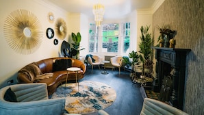 A living room with a brown leather sofa, a round coffee table, and a fireplace. There are two chairs, a large mirror, and a chandelier. The room has a large window with a view of greenery outside. - Cranleigh Boutique (Windermere)