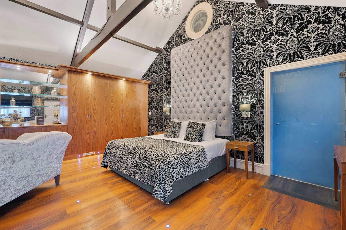 A bedroom with a tufted headboard, a bed with a patterned bedspread, a wooden floor, a blue door, and a decorative wall with a floral pattern.