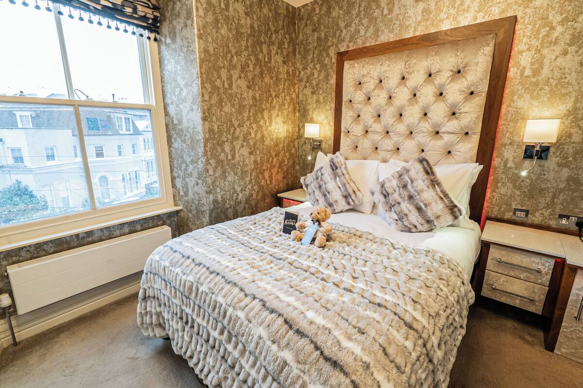 A hotel room with a large bed, a tufted headboard, two bedside lamps, a window with a view of buildings, and a radiator under the window.