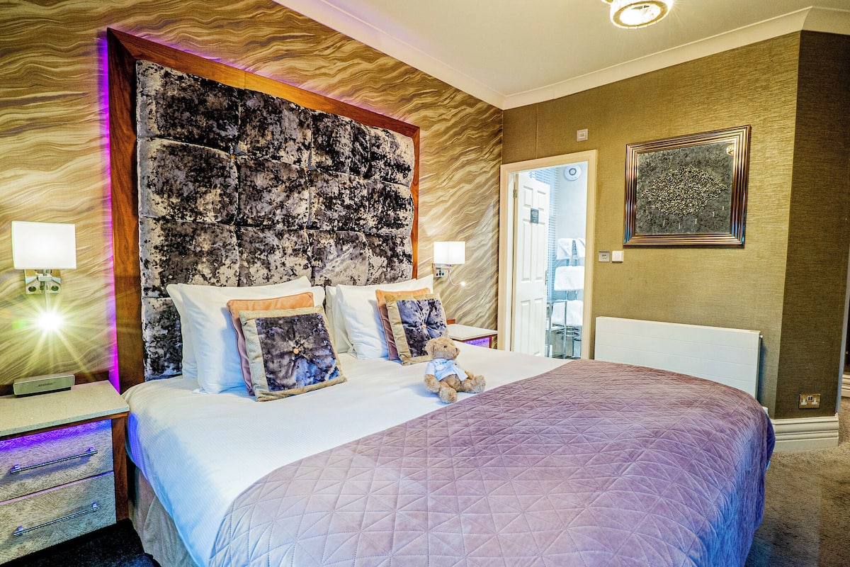 A neatly made bed with a quilted cover, a plush headboard, and a teddy bear on the pillows.
