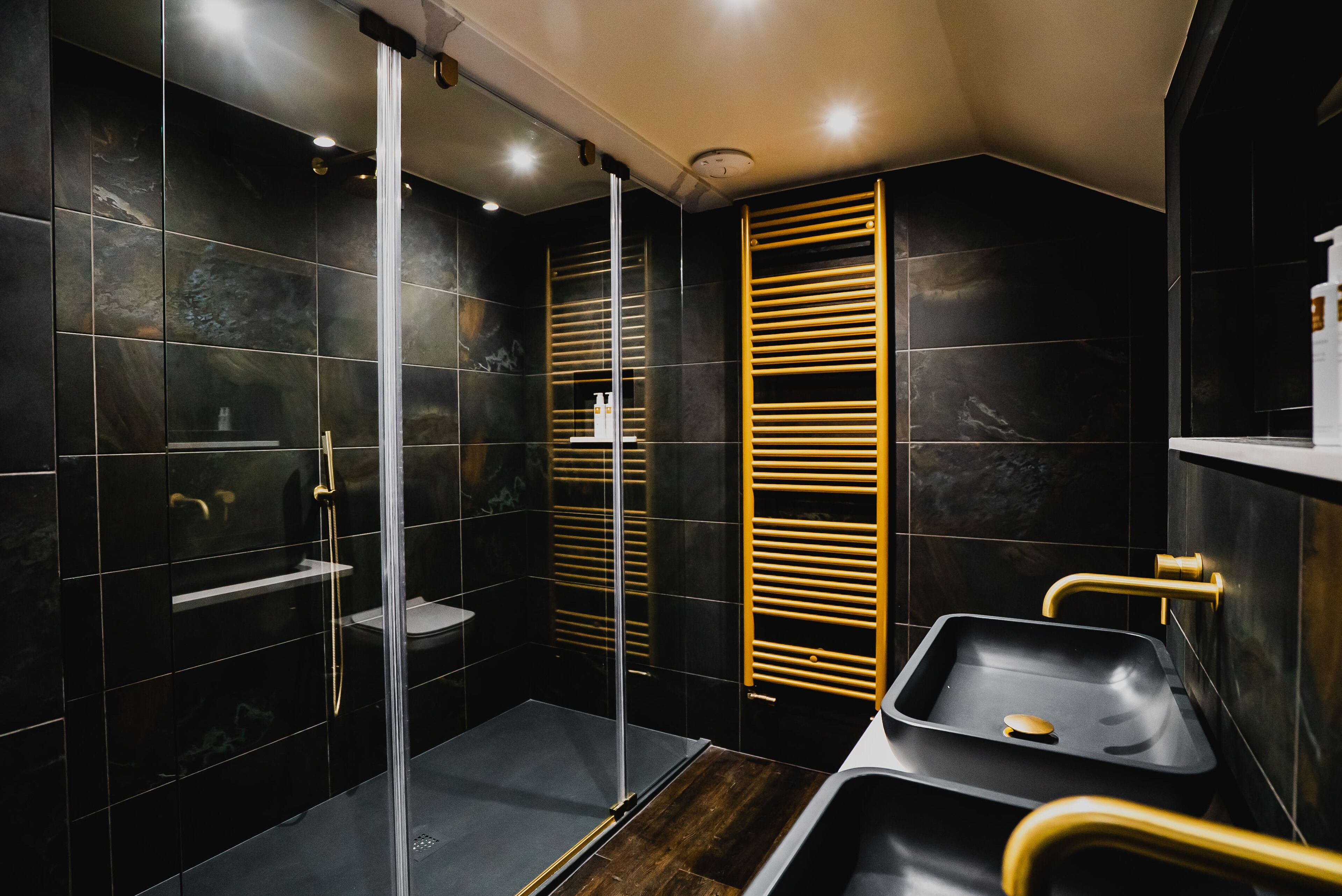 A modern bathroom with a glass shower enclosure, a wooden towel rack, and a black sink with gold fixtures.