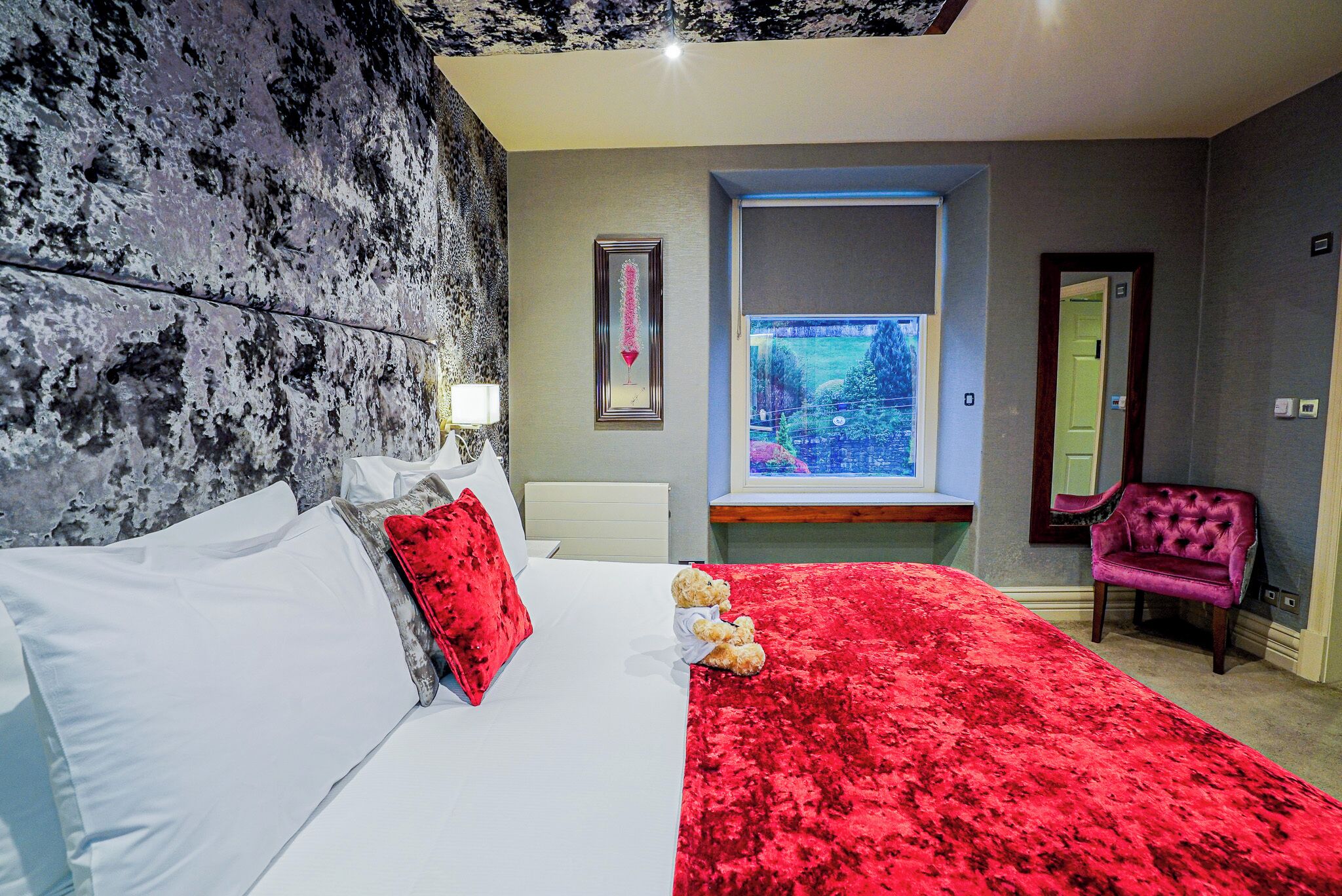 Une chambre avec un grand lit, un plaid rouge, un ours en peluche, une fenêtre donnant sur un beau paysage, un miroir et une chaise violette.