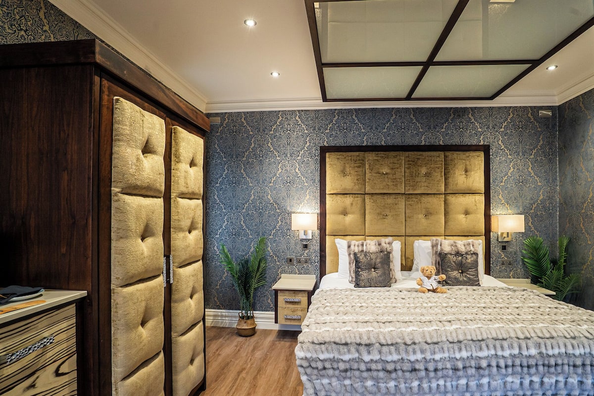 A bedroom with a large bed, a tufted headboard, two bedside lamps, and a nightstand with a plant.