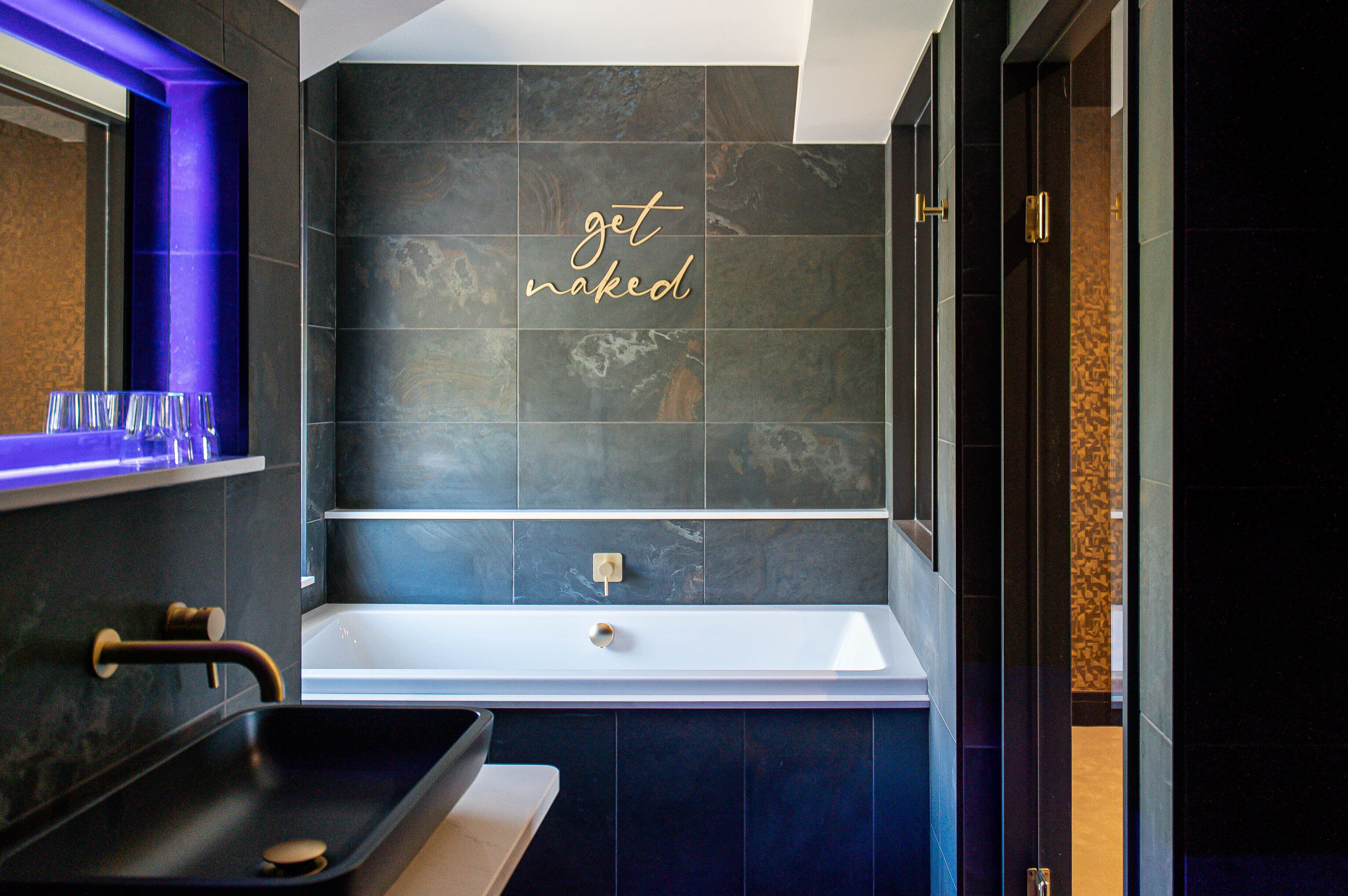A modern bathroom with a large bathtub, a vanity with a sink, and a mirror. The wall features a 'get naked' sign.