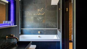 A modern bathroom with a large bathtub, a vanity with a sink, and a mirror. The wall features a 'get naked' sign.