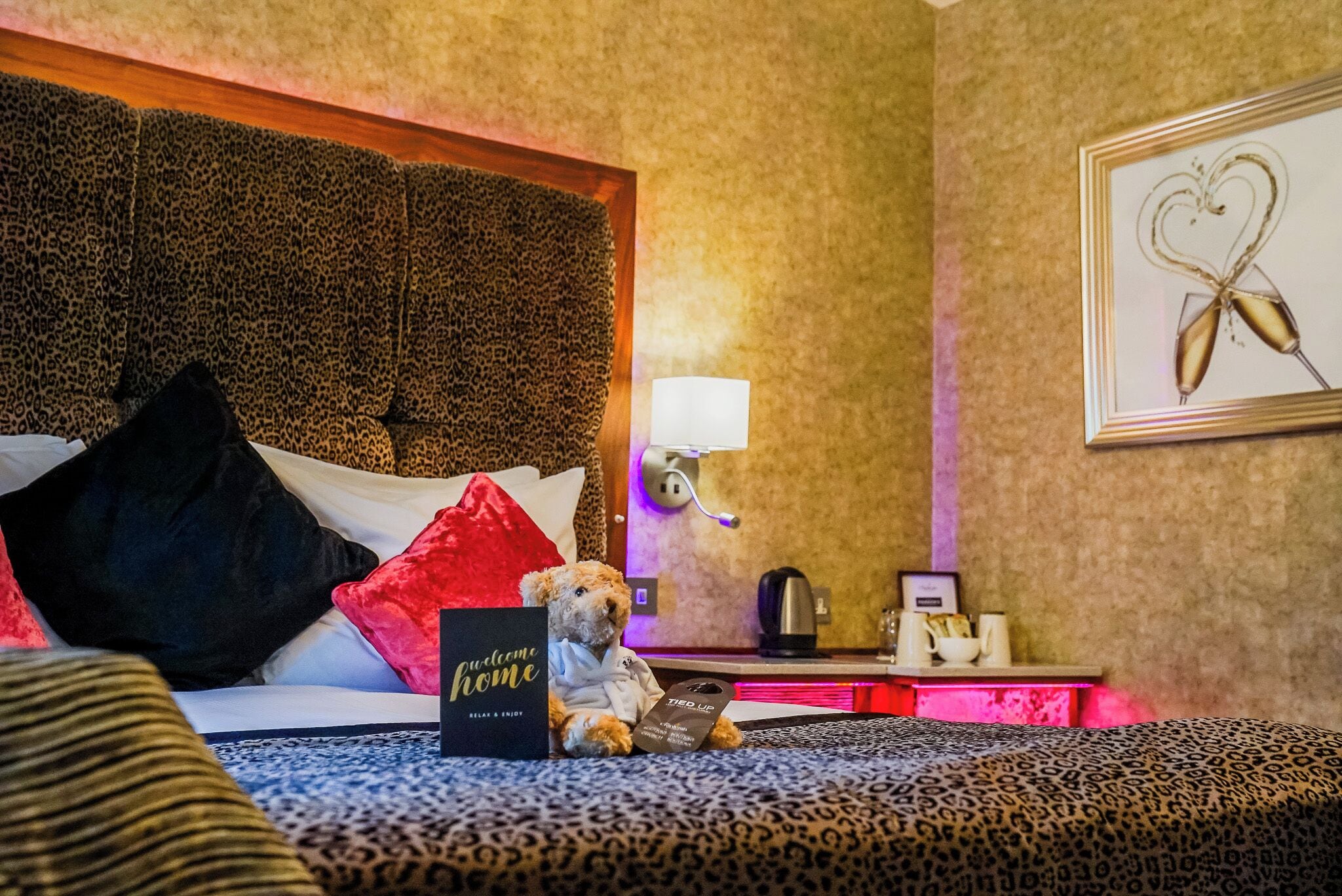 A hotel room with a leopard print bedspread, a plush teddy bear, a welcome sign, and a framed picture on the wall.