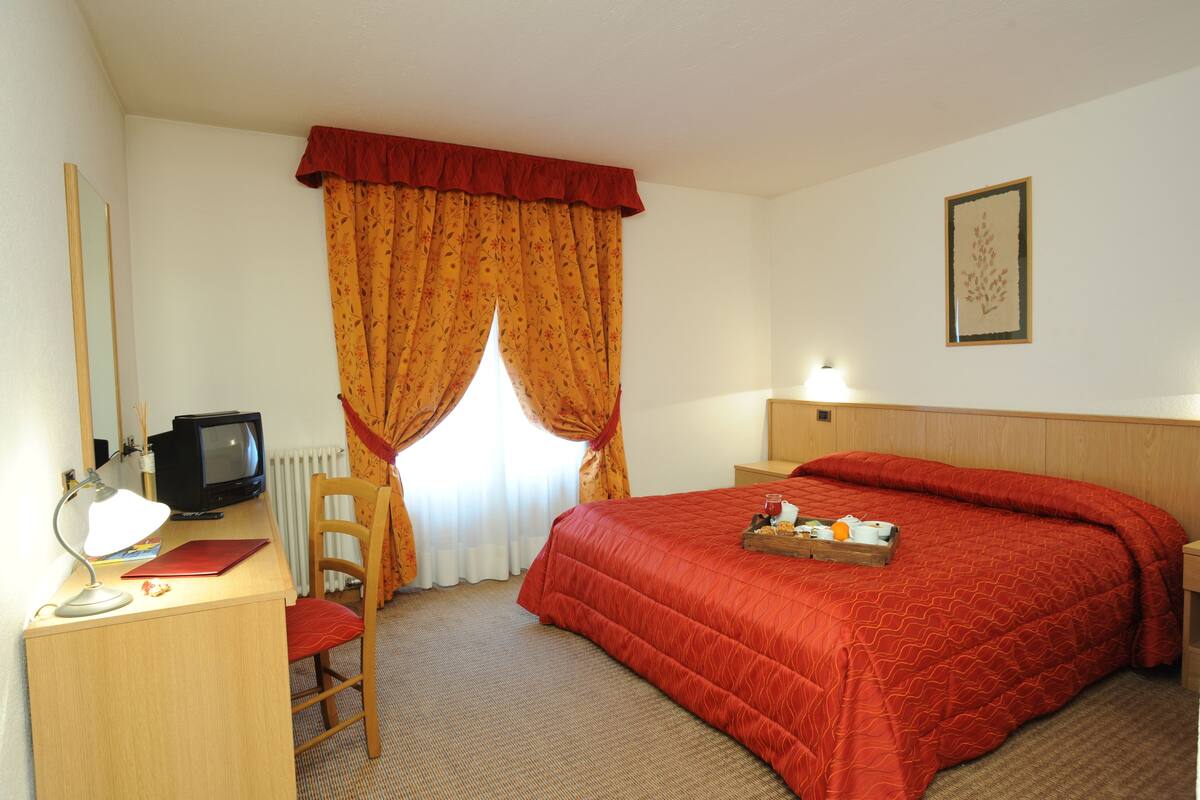 A hotel room with a bed, a desk with a computer and lamp, a chair, a window with curtains, and a framed picture on the wall.