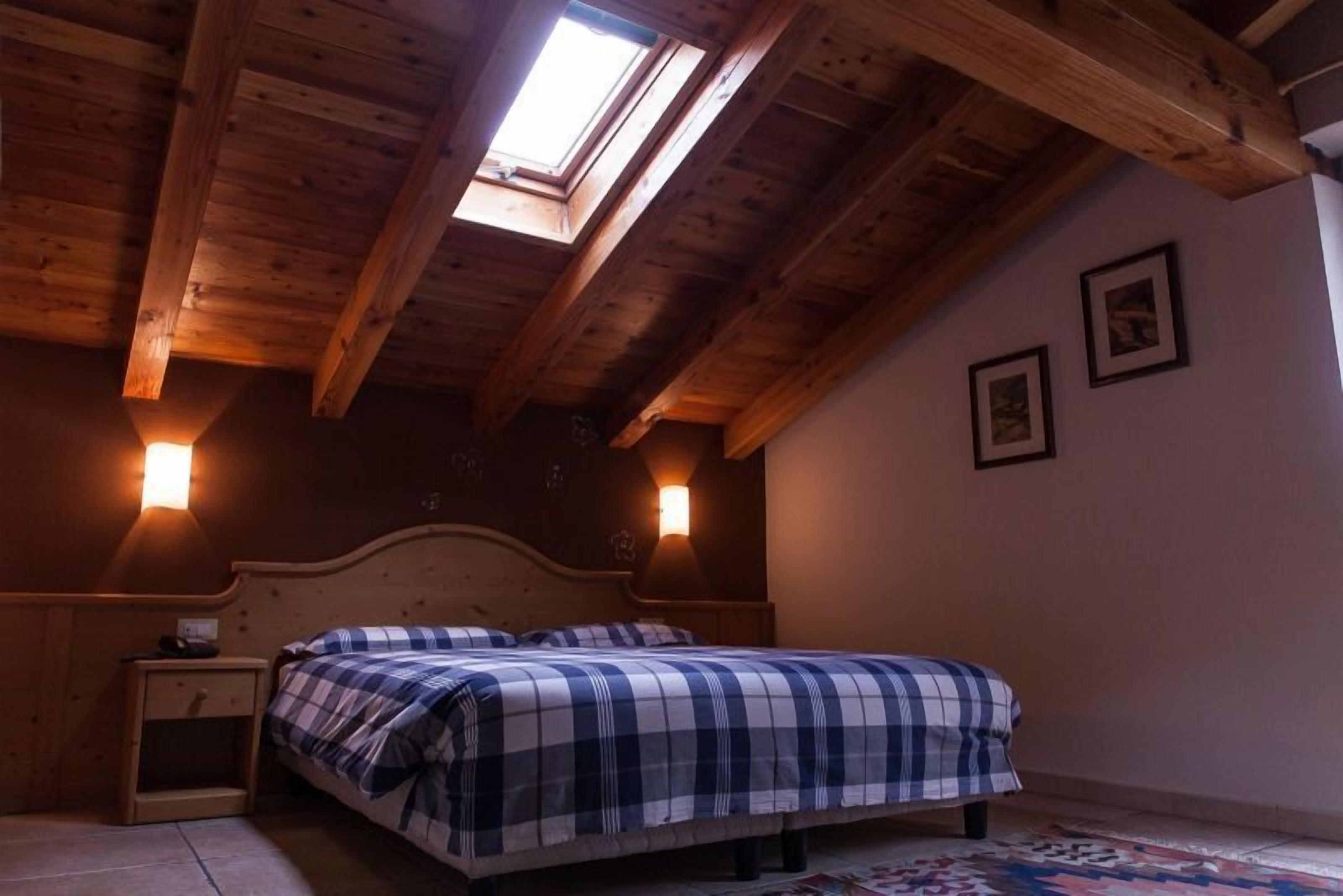 A bedroom with a sloped ceiling, wooden beams, a skylight, two bedside tables with lamps, a bed with a checkered bedspread, and two framed pictures on the wall.