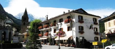 Un hotel con facciata bianca, diversi balconi fioriti e uno sfondo montuoso.
