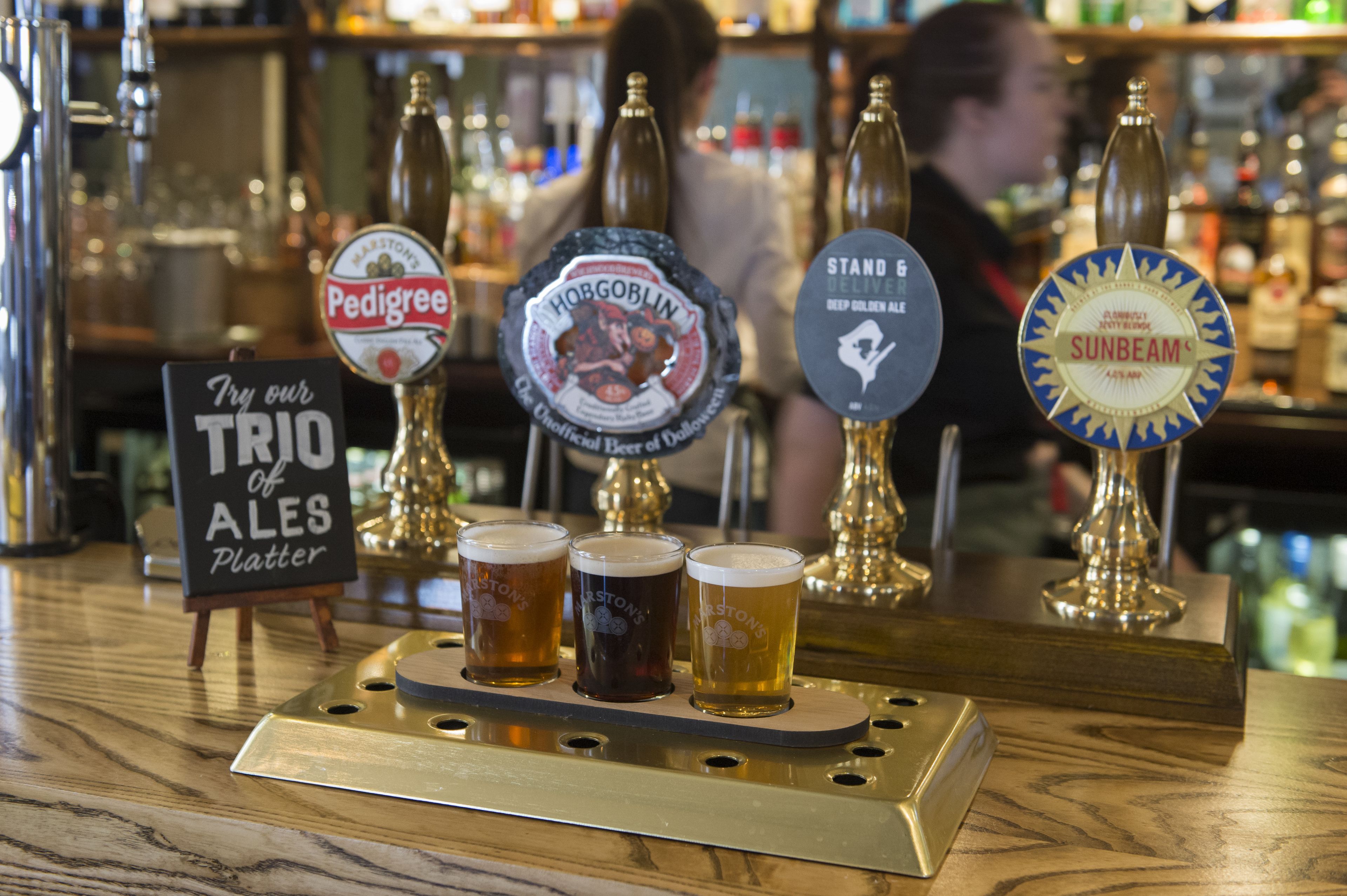 A bar counter with four beer taps and a sign offering a trio of ales.