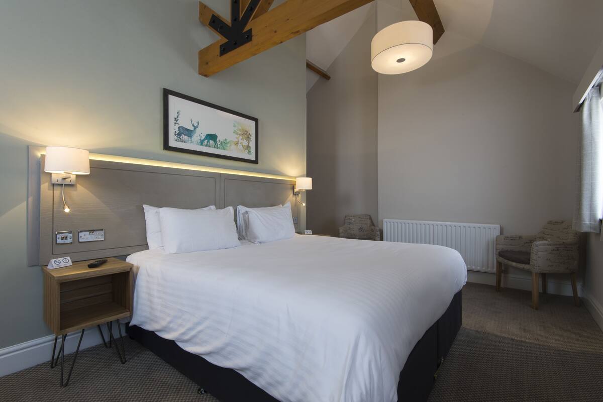 Un cuarto de hotel ordenado con una cama grande, lámparas de noche, un cuadro en la pared y una silla junto a la ventana.