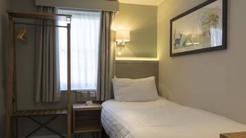 Een hotelkamer met een bed, nachtkastje, lamp, raam met gordijnen en een schilderij aan de muur.