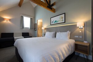 Quarto de hotel com uma cama grande, mesa de cabeceira, cortinas em janela, quadro na parede e teto com vigas aparentes.