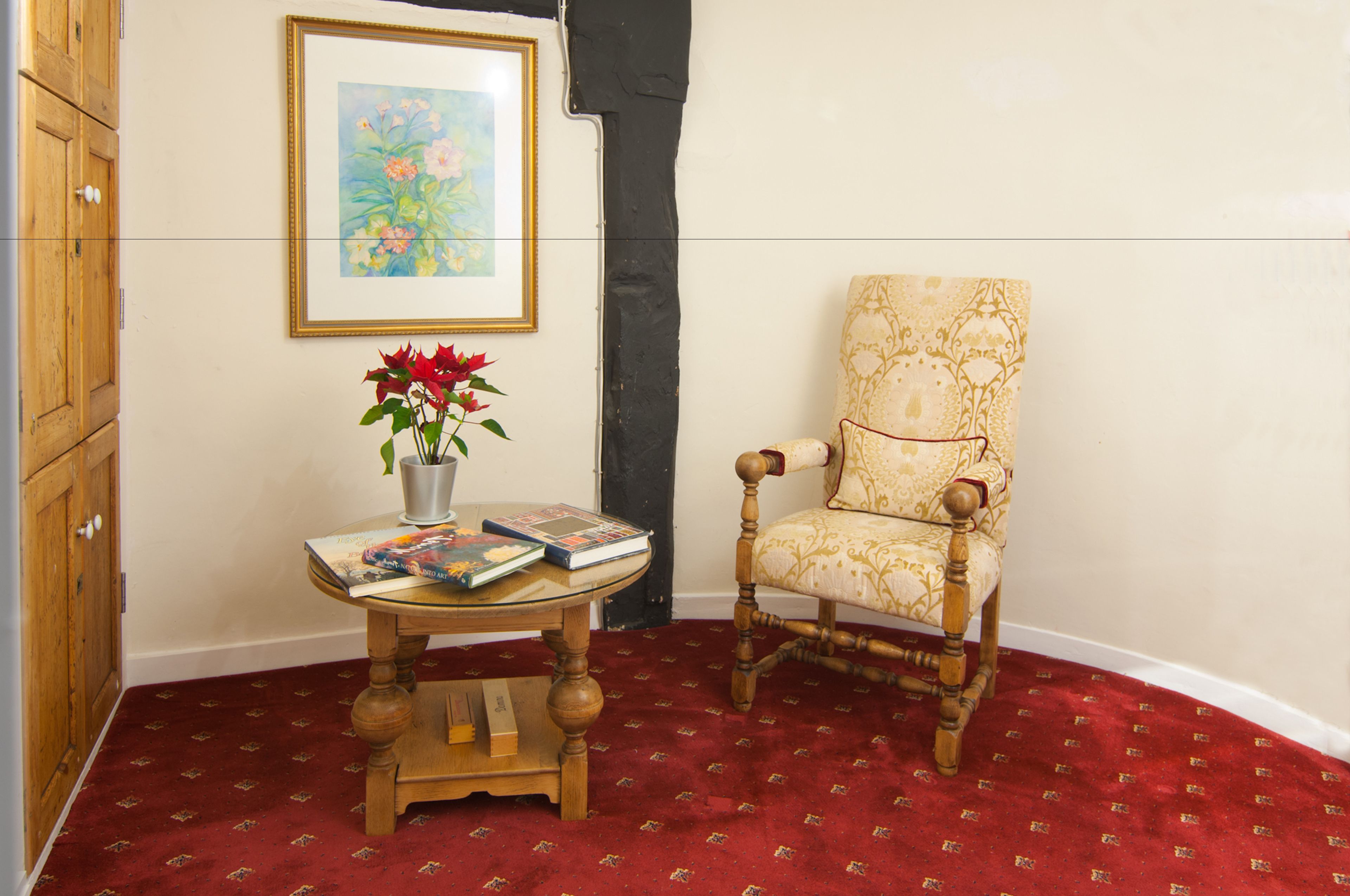 Una stanza con una sedia in legno, un tavolino rotondo con un vaso di fiori e un quadro appeso al muro.