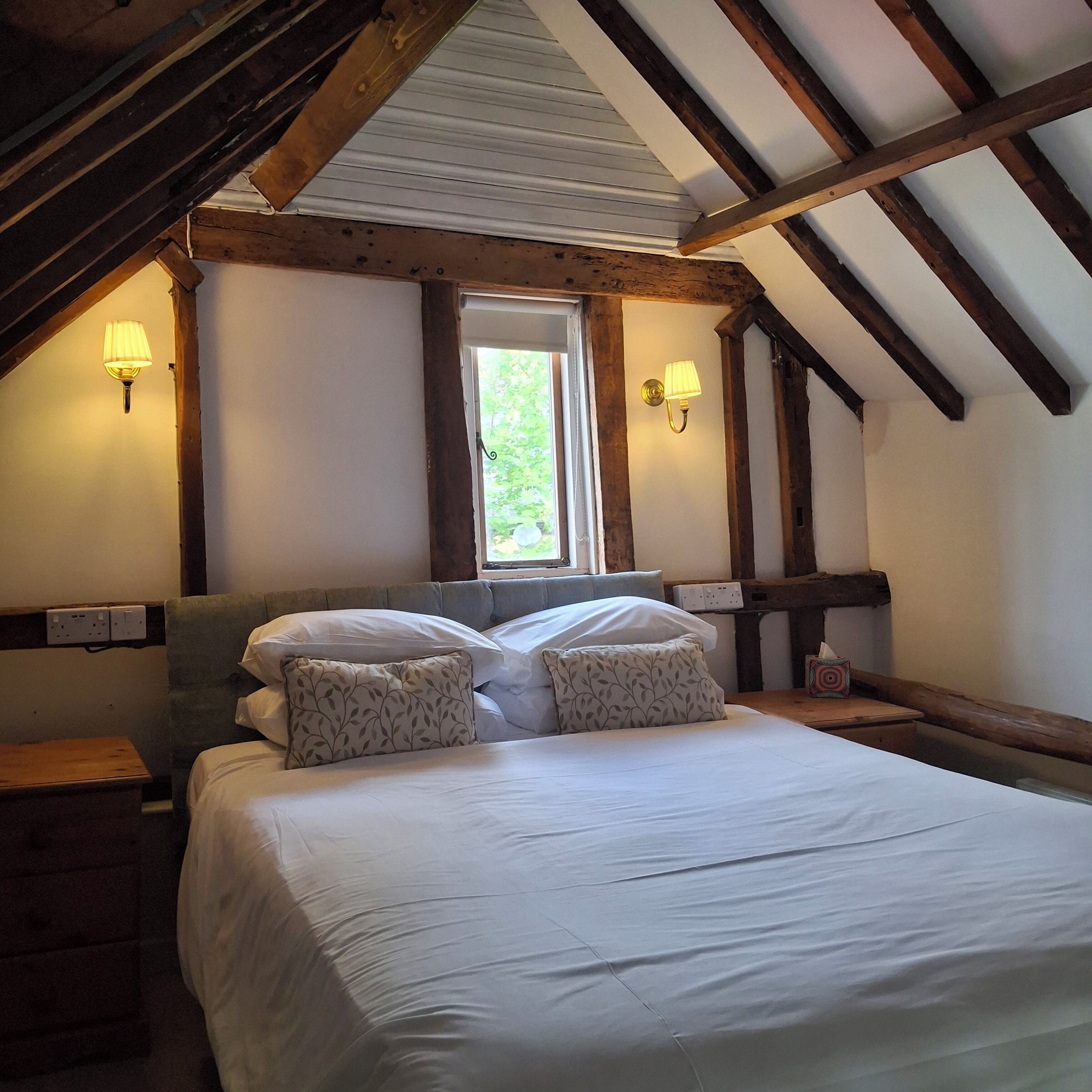 Un dormitorio con techo inclinado, una cama con ropa de cama blanca, dos lámparas fijadas a la pared y un ventanal con vistas a zonas verdes.