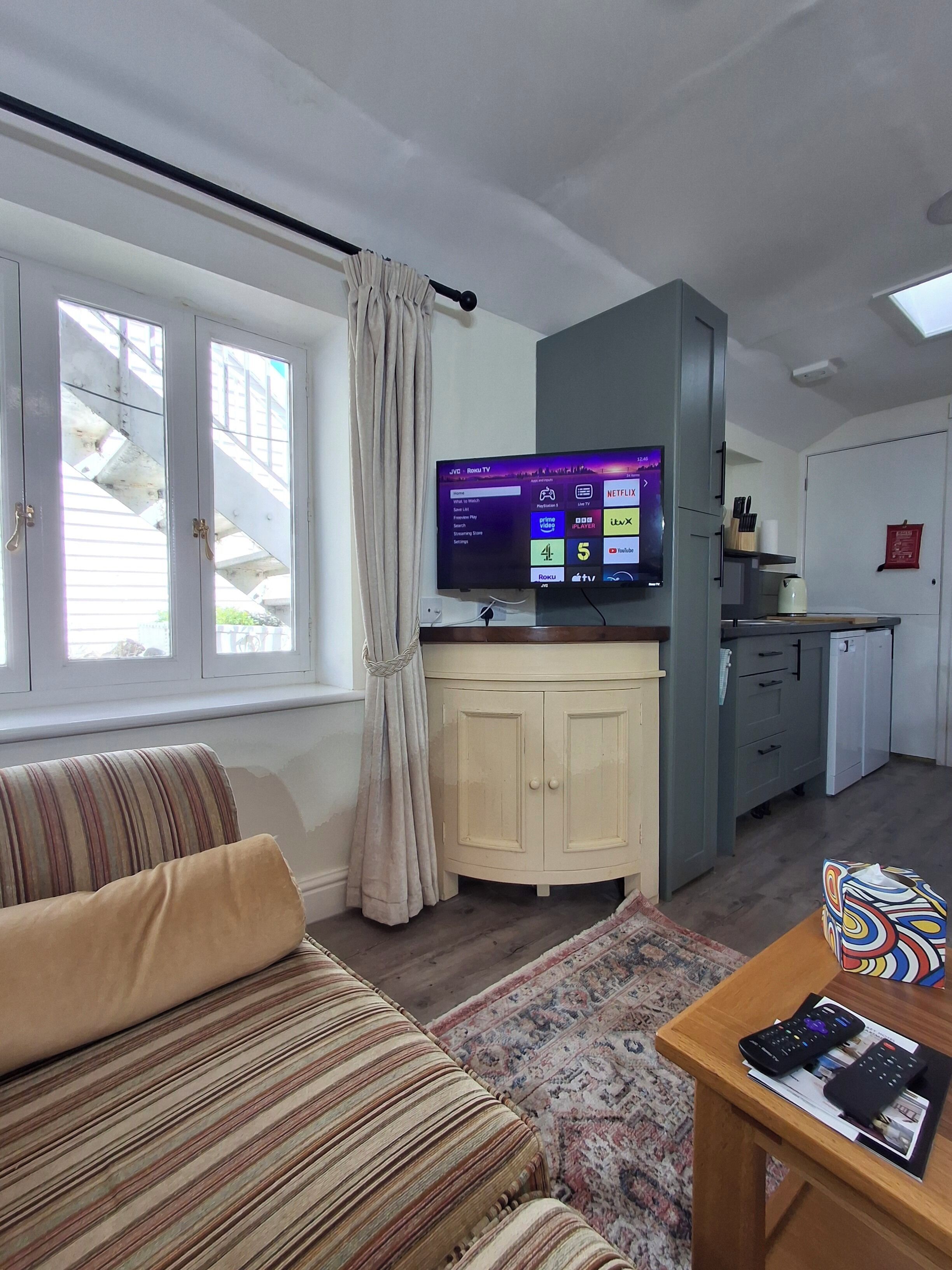 A living room with a sofa, a coffee table with remote controls, a television displaying a streaming service interface, a kitchen area with cabinets, and a window with curtains.