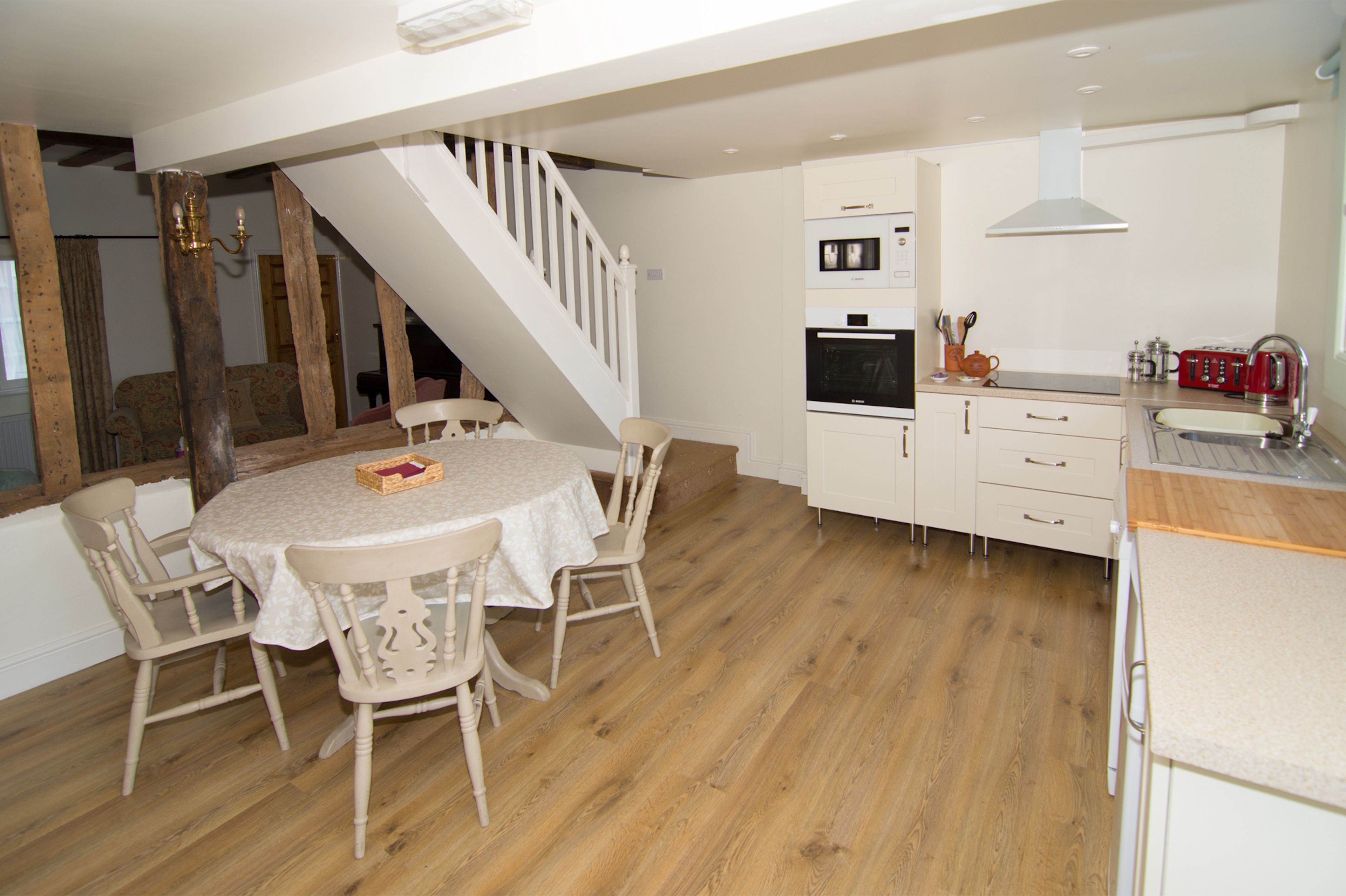 Un coin repas avec une table ronde et des chaises, une cuisine avec des meubles blancs et des appareils électroménagers, et un escalier en bois.