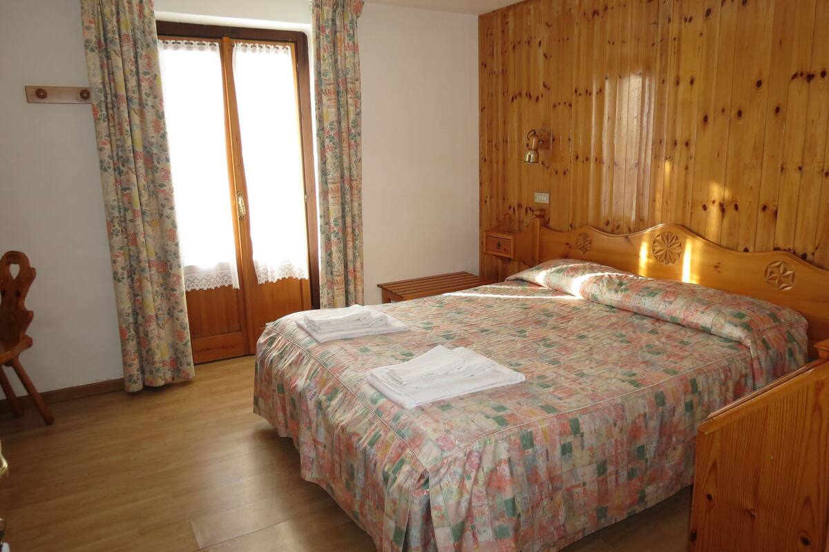 Une chambre à coucher avec une tête de lit en bois, un lit recouvert d’une literie à motifs floraux, une table de chevet en bois et une chaise en bois.
