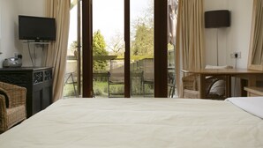 Un dormitorio con una cama grande, televisión, escritorio y una silla. Hay un balcón con vistas a árboles y una valla.