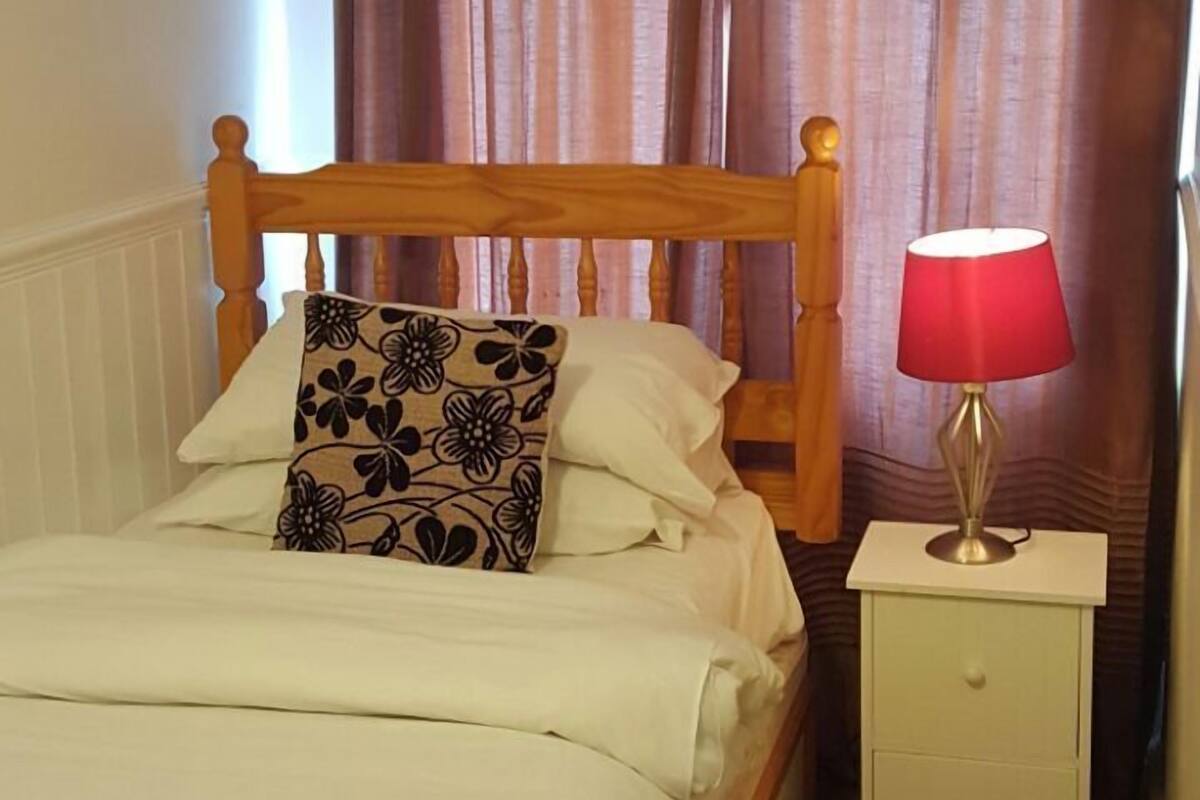 A bedroom with a wooden bed, white bedding, a floral pillow, a nightstand with a lamp, and brown curtains.