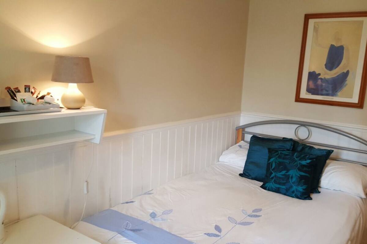 A bedroom with a bed, a bedside table, a lamp, a shelf with books and a framed picture.