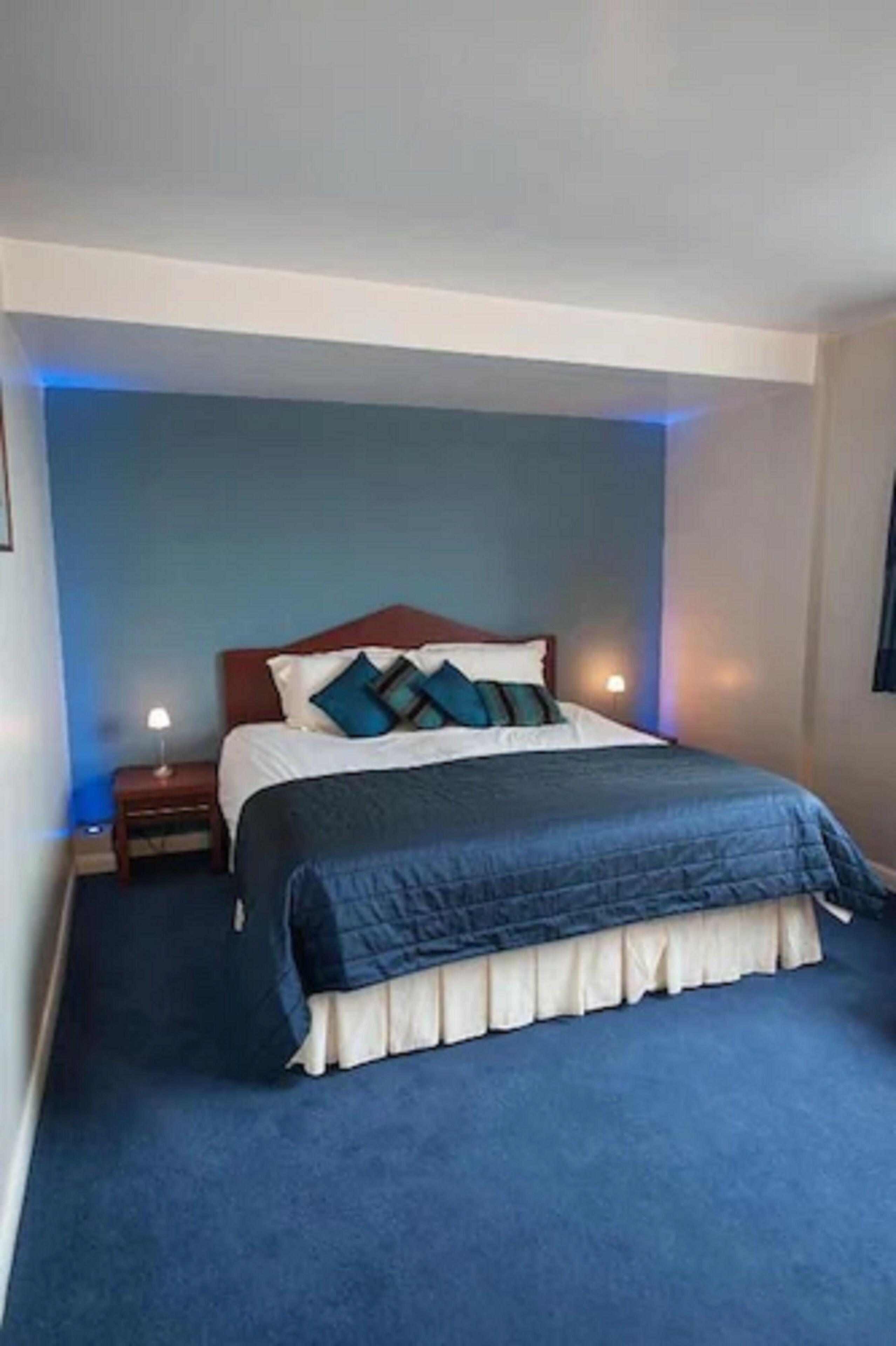A neatly made bed with a blue bedspread and white bedding, a wooden headboard, and a small bedside table with a lamp.
